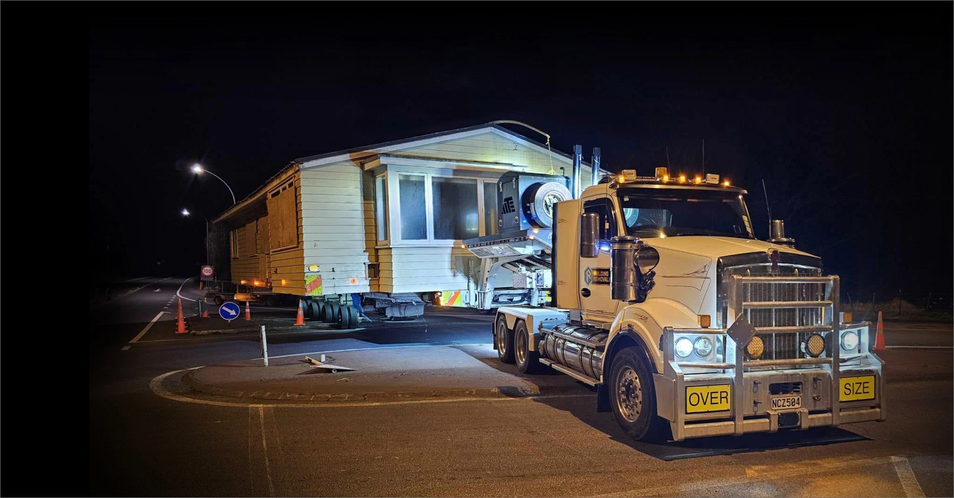 A weatherboard house on a hydraulic house trailer at night, lit by street lights and the truck's beacons, with oversize load boards fitted and traffic cones set out